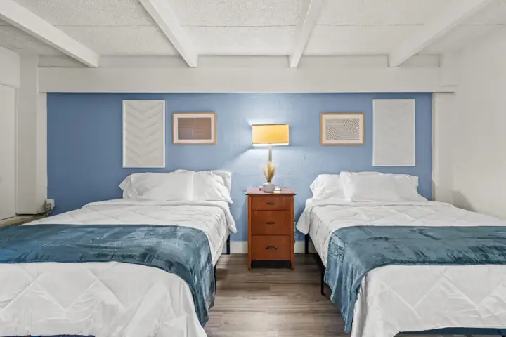 A bedroom with two double beds separated by a wooden nightstand, blue accent wall, framed artwork, and a table lamp