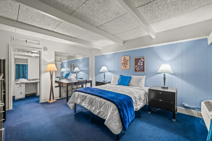 A hotel room with a bed covered in white bedding and blue accents, a black nightstand with a lamp, and blue wall paneling