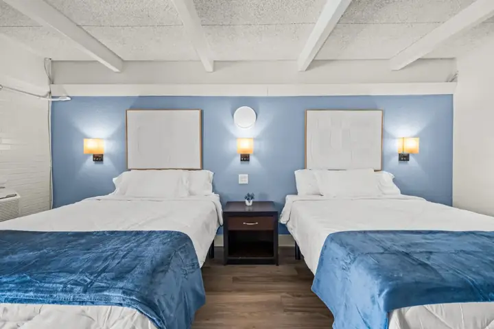 A hotel room with two beds, each with blue blankets, white sheets, and pillows. There is a nightstand between the beds