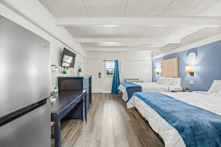 A hotel room with two beds, one with a blue blanket, a small nightstand, and wall-mounted lamps, with a blue accent wall