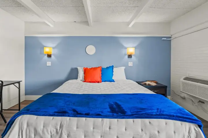 A neatly made bed with white bedding and two colorful pillows, one red and one blue, against a blue accent wall with two