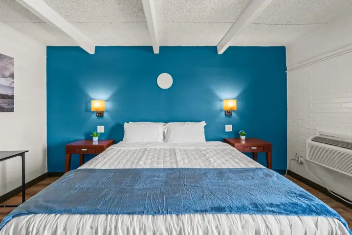 A neatly made bed with white pillows and a white comforter, has a blue blanket at the foot, in a room with a blue accent