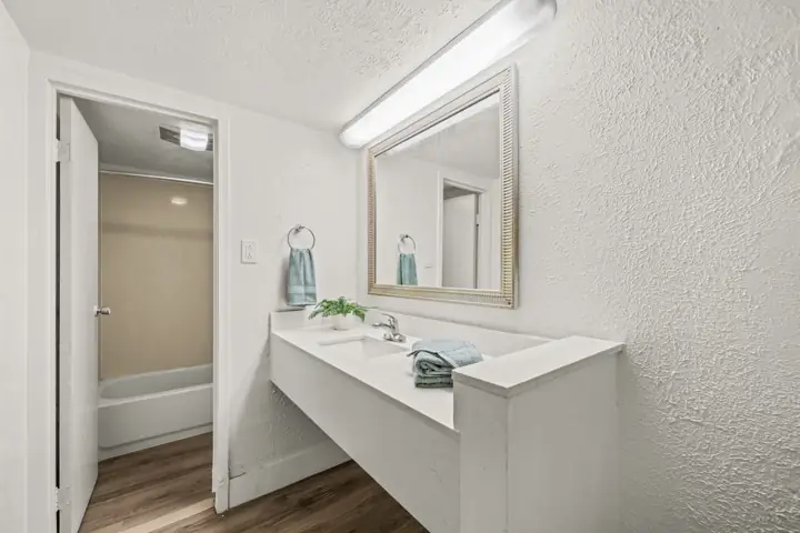A small bathroom with a white vanity, large mirror, and a bathtub/shower combination. The room has white textured walls