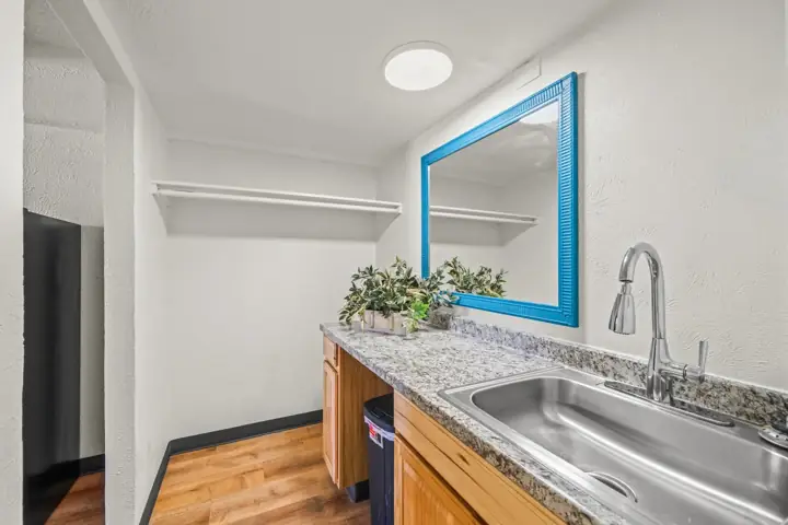A small kitchenette with a granite countertop, wooden cabinets, a stainless steel sink, a blue framed mirror, a potted