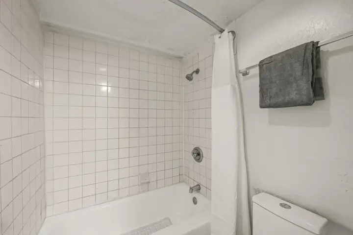 Bathroom with a tiled shower area, shower head, white tub, towel rod with a gray towel, and part of a toilet