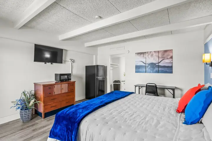 Hotel room with a bed, blue and red pillows, a blue blanket, a wall-mounted TV, a wooden dresser, a microwave, a