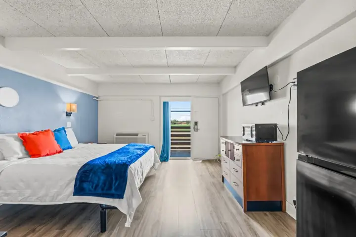 Hotel room with a bed featuring red and blue pillows, a blue throw blanket, a wooden dresser with a microwave and a small
