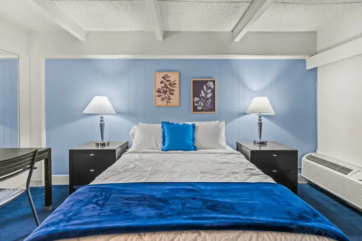 Hotel room with a bed, two nightstands with lamps, artwork above the bed, dark furniture, blue accents, and a window air