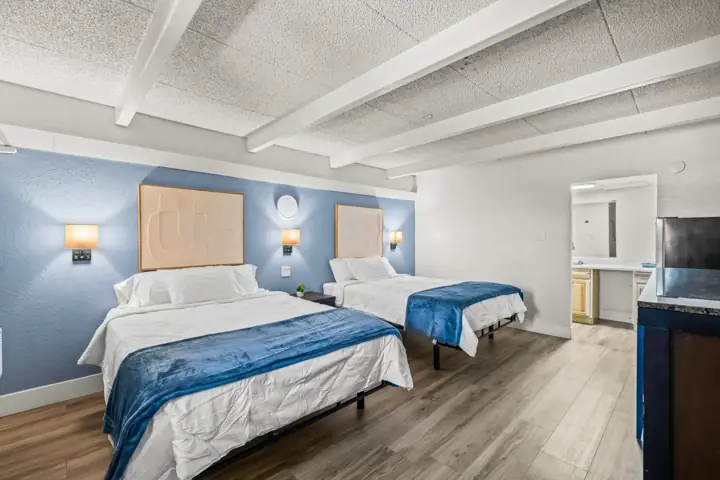 Hotel room with two beds with white linens and blue throws, blue accent wall, and wooden floors. There are wall-mounted