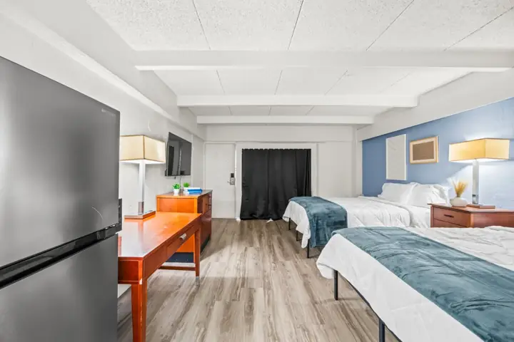 Interior of a hotel room with two beds, a flat-screen TV on a wooden dresser, a refrigerator, a small table, and