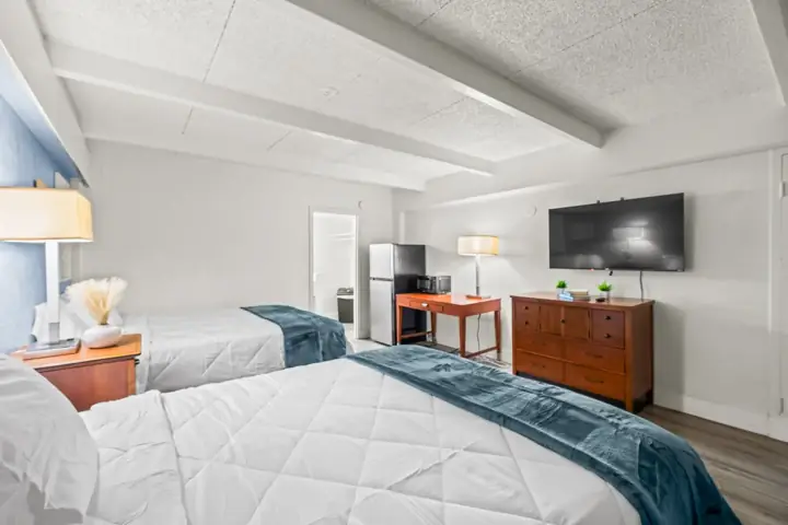 Two beds in a room with white quilted bedding and blue throws, a wooden nightstand with a lamp and decorative vase, a